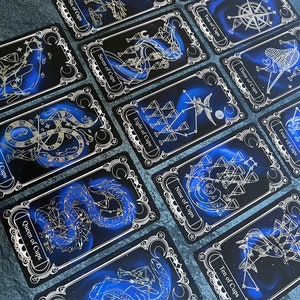May include: A collection of tarot cards from the suit of Cups, featuring dark blue backgrounds with gold foil constellation-style illustrations. Each card displays a specific title, such as Ace of Cups, Six of Cups, and King of Cups, framed by ornate borders.