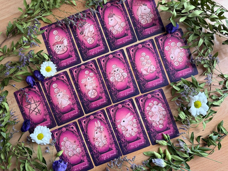 May include: A collection of fourteen tarot cards featuring pink and purple backgrounds with illustrations of white rabbits and pentacles. Each card is framed in an ornate, dark border and displays a unique scene with rabbits interacting with coins.