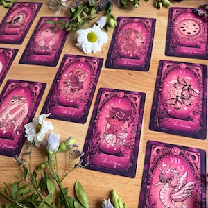 May include: A collection of rectangular tarot cards featuring pink and dark purple illustrations of animals and mystical symbols. Each card displays intricate borders and text labels such as The Magician, The Fool, and The Empress, arranged on a wooden surface with scattered white flowers.