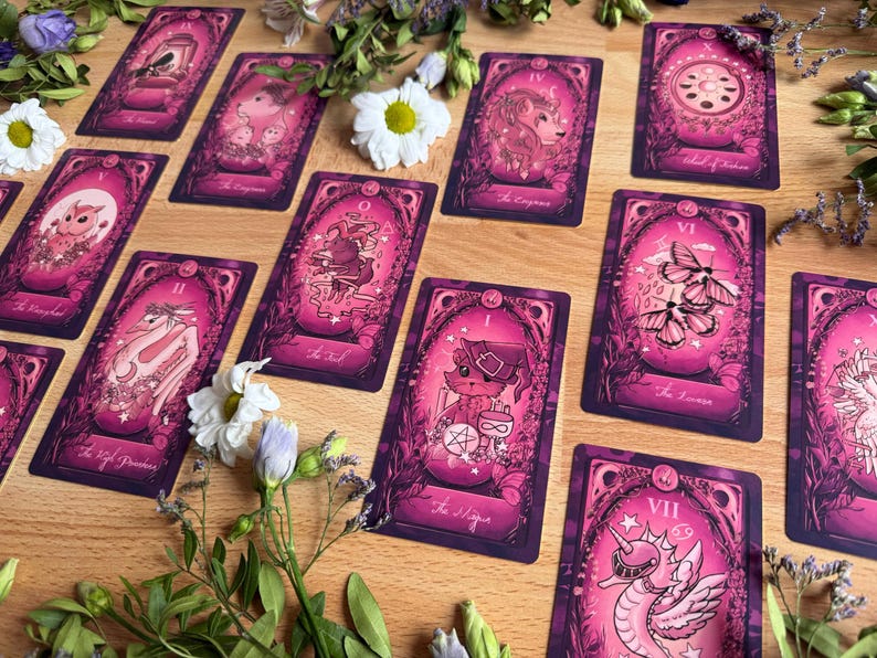 May include: A collection of rectangular tarot cards featuring pink and dark purple illustrations of animals and mystical symbols. Each card displays intricate borders and text labels such as The Magician, The Fool, and The Empress, arranged on a wooden surface with scattered white flowers.