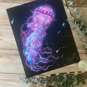 May include: A vibrant art print featuring a jellyfish illustration. The jellyfish is depicted in shades of pink, purple, and blue against a dark background. The tentacles are long and flowing, and the overall design has a dreamy, ethereal quality.