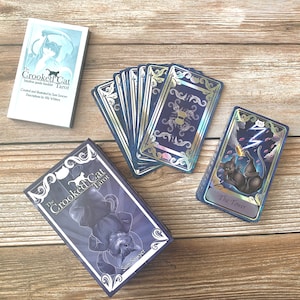 May include: A tarot card deck titled The Crooked Cat Tarot, featuring a guide booklet, a box with a black cat illustration, and cards with dark blue, silver, and holographic accents. One card displays The Tower with a black cat and lightning.