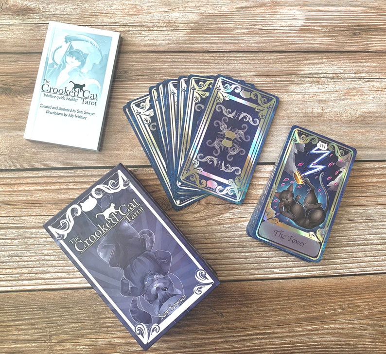 May include: A tarot card deck titled The Crooked Cat Tarot, featuring a guide booklet, a box with a black cat illustration, and cards with dark blue, silver, and holographic accents. One card displays The Tower with a black cat and lightning.
