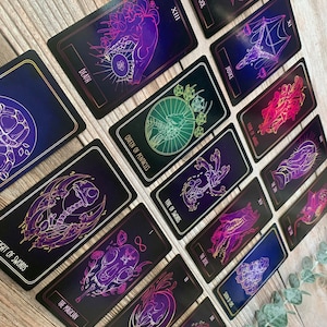 May include: A collection of tarot cards with black backgrounds and vibrant neon illustrations. The cards feature images of skulls, hands, and other mystical symbols, with titles like "Death," "Justice," and "The Fool." The cards are arranged on a wooden surface.