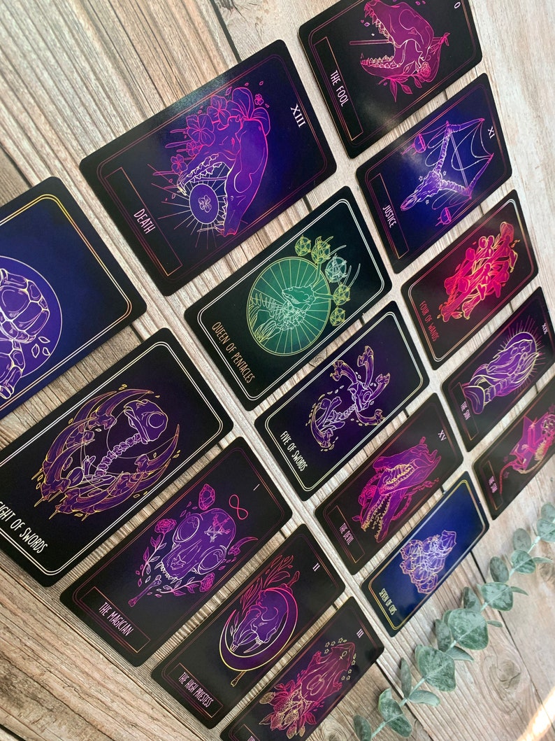 May include: A collection of tarot cards with black backgrounds and vibrant neon illustrations. The cards feature images of skulls, hands, and other mystical symbols, with titles like "Death," "Justice," and "The Fool." The cards are arranged on a wooden surface.