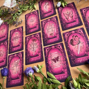 May include: A collection of tarot cards featuring a pink and purple color palette with intricate illustrations of swords, butterflies, and moths. Each card is framed by ornate borders and surrounded by fresh white daisies and purple flowers.