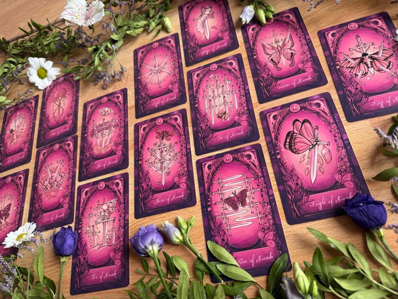 May include: A collection of tarot cards featuring a pink and purple color palette with intricate illustrations of swords, butterflies, and moths. Each card is framed by ornate borders and surrounded by fresh white daisies and purple flowers.