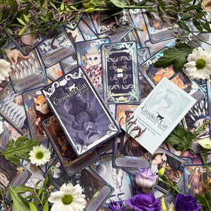 May include: A deck of The Crooked Cat Tarot cards featuring purple and silver holographic accents, a matching box with a cat illustration, and an intuitive guide booklet, all surrounded by white daisies, purple roses, and green foliage.