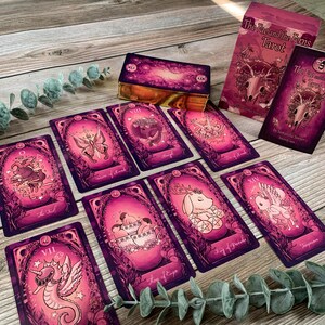 May include: A deck of The Fae and the Ferns Tarot cards featuring vibrant magenta and pink illustrations of animals and mythical creatures. The set includes a box, a guidebook, and several individual cards displayed with gold-gilded edges.