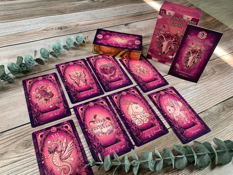 May include: A deck of The Fae and the Ferns Tarot cards featuring vibrant magenta and pink illustrations of animals and mythical creatures. The set includes a box, a guidebook, and several individual cards displayed with gold-gilded edges.