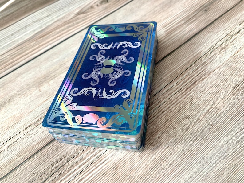 May include: A rectangular deck of cards featuring a deep blue background with a holographic finish. The card back displays a symmetrical design with intricate silver filigree patterns, swirling motifs, and a central emblem of a cat silhouette.