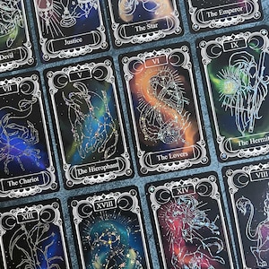 May include: A collection of rectangular tarot cards featuring black backgrounds with colorful nebula-like patterns and white line art illustrations. Each card displays a Roman numeral, a title such as Justice, The Star, The Emperor, The Hierophant, The Lovers, The Hermit, The Chariot, Death, The Moon, and Temperance.