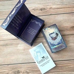 May include: A deck of The Crooked Cat Tarot cards with a holographic Ace of Cups card on top, a matching purple storage box with printed text, and an intuitive guide booklet. The box interior features text about an adventure and a paw-shaped symbol.