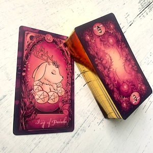 May include: A tarot card with a pink and gold design. The card features a white, furry creature with antlers and closed eyes. The creature is sitting on a bed of pink and gold coins. The card reads "King of Pentacles".