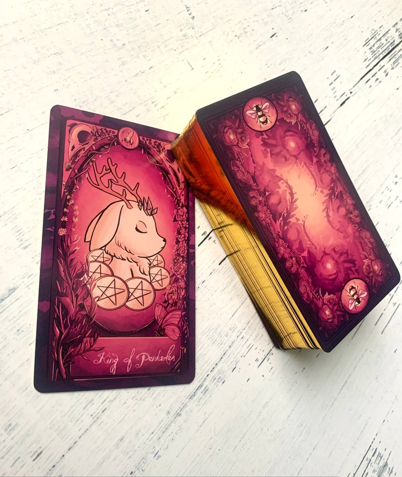 May include: A tarot card with a pink and gold design. The card features a white, furry creature with antlers and closed eyes. The creature is sitting on a bed of pink and gold coins. The card reads "King of Pentacles".