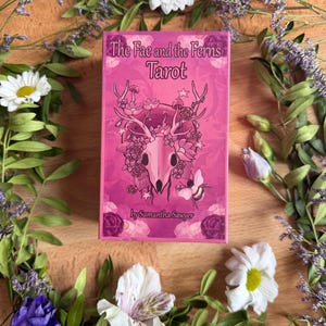 May include: A rectangular tarot deck box in shades of pink featuring the text The Fae and the Ferns Tarot by Samantha Sawyer. The front illustration depicts a deer skull adorned with flowers, mushrooms, and a bee, surrounded by floral motifs.