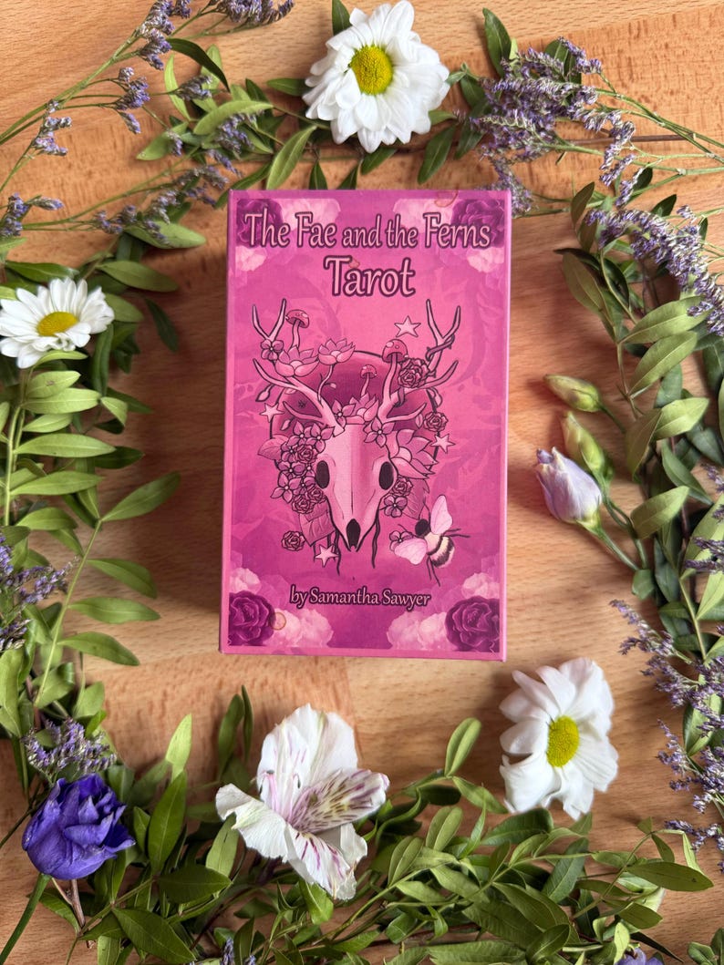 May include: A rectangular tarot deck box in shades of pink featuring the text The Fae and the Ferns Tarot by Samantha Sawyer. The front illustration depicts a deer skull adorned with flowers, mushrooms, and a bee, surrounded by floral motifs.