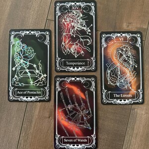 May include: Four rectangular tarot cards with black backgrounds and white line art. The cards are labeled Ace of Pentacles, Temperance, The Lovers, and Seven of Wands. Each card features a unique celestial or symbolic illustration with holographic accents.