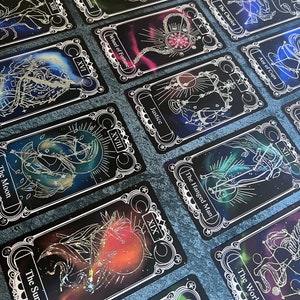 May include: A collection of tarot cards featuring black backgrounds with colorful nebula-like patterns and silver foil line art illustrations. Each card displays a Roman numeral and a title such as The Sun, The Moon, The Hermit, and The World.