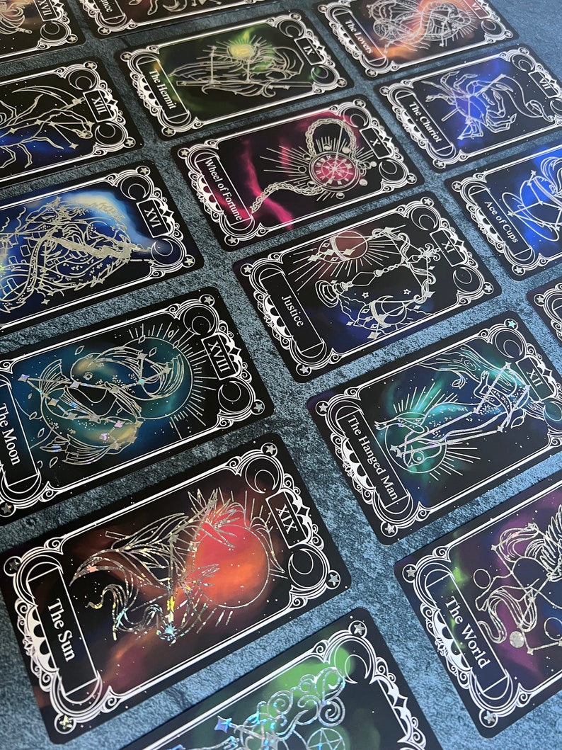May include: A collection of tarot cards featuring black backgrounds with colorful nebula-like patterns and silver foil line art illustrations. Each card displays a Roman numeral and a title such as The Sun, The Moon, The Hermit, and The World.