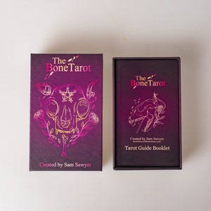 May include: A tarot card set with a dark purple box and a matching booklet. The box features gold foil illustrations of a skull, roses, and the text "The Bone Tarot." The booklet reads "Tarot Guide Booklet."