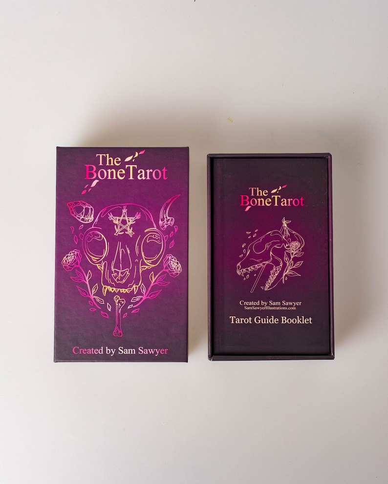 May include: A tarot card set with a dark purple box and a matching booklet. The box features gold foil illustrations of a skull, roses, and the text "The Bone Tarot." The booklet reads "Tarot Guide Booklet."