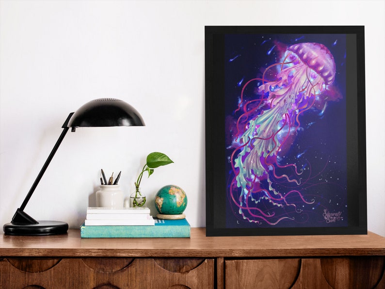 May include: A framed art print featuring a vibrant jellyfish illustration. The artwork showcases a jellyfish with a pink, purple, and teal color palette against a dark blue background. The print is displayed on a wooden surface, alongside a black desk lamp, books, and a globe.
