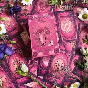 May include: A rectangular tarot deck box titled The Fae and the Ferns Tarot by Samantha Sawyer, featuring a pink illustration of a deer skull adorned with flowers and a bee, surrounded by multiple tarot cards with matching pink artwork.