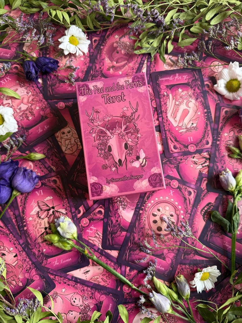 May include: A rectangular tarot deck box titled The Fae and the Ferns Tarot by Samantha Sawyer, featuring a pink illustration of a deer skull adorned with flowers and a bee, surrounded by multiple tarot cards with matching pink artwork.