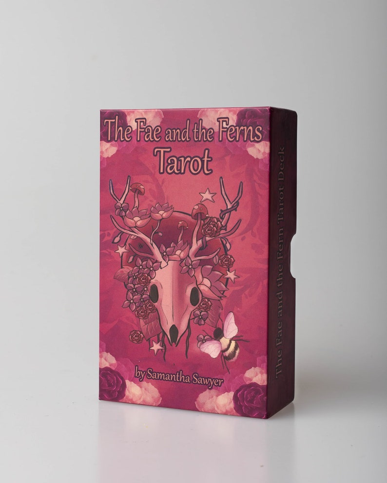 May include: A pink box with a skull and floral design. The box is for a tarot deck called "The Fae and the Ferns Tarot" by Samantha Sawyer.