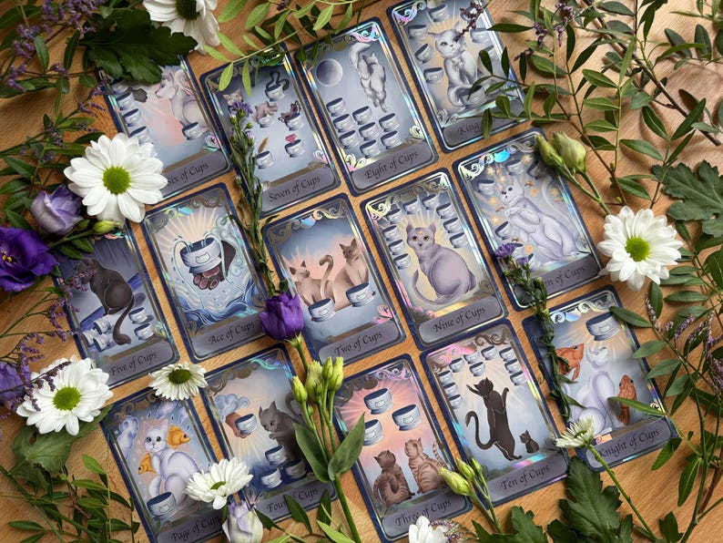 May include: A collection of tarot cards from the suit of cups, each featuring illustrations of cats and cups, arranged on a wooden surface surrounded by white daisies and purple flowers. The cards include labels like Ace of Cups and Ten of Cups.