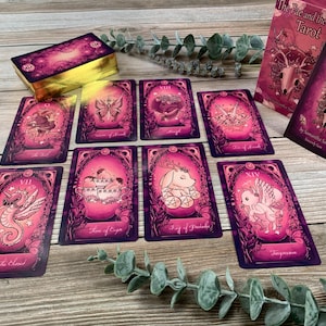 May include: A deck of The Fae and the Ferns Tarot cards with a pink and purple color palette. The cards feature illustrations of creatures and nature motifs. A box with the title The Fae and the Ferns Tarot by Samantha Sawyer is included.