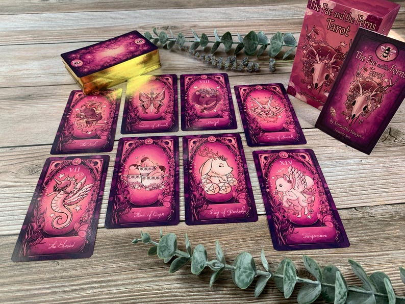 May include: A deck of The Fae and the Ferns Tarot cards with a pink and purple color palette. The cards feature illustrations of creatures and nature motifs. A box with the title The Fae and the Ferns Tarot by Samantha Sawyer is included.