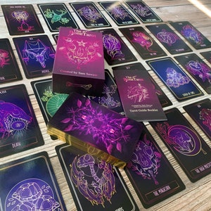 May include: A tarot deck with a box and guidebook. The cards feature dark backgrounds with purple and gold illustrations of skulls, flowers, and other symbols. The box and guidebook read "The Bone Tarot" and "Created by Sam Sawyer."