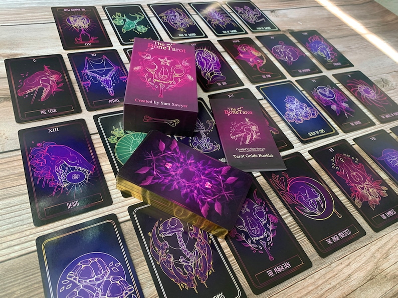 May include: A tarot deck with a box and guidebook. The cards feature dark backgrounds with purple and gold illustrations of skulls, flowers, and other symbols. The box and guidebook read "The Bone Tarot" and "Created by Sam Sawyer."