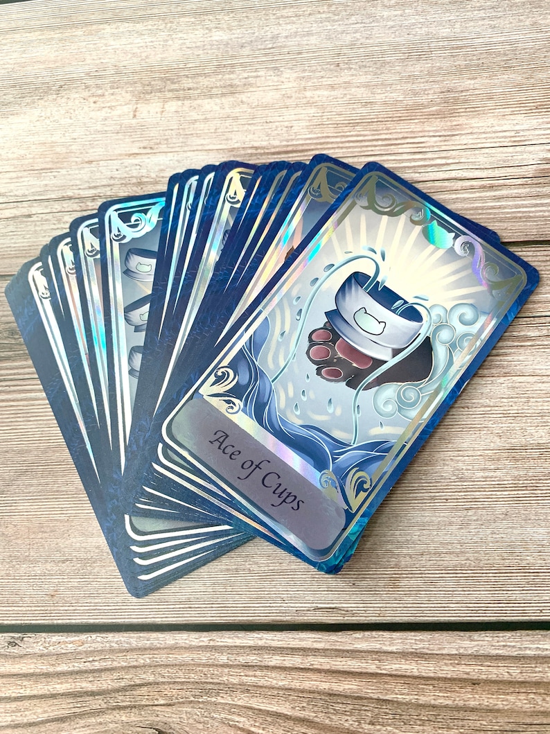 May include: A fan of tarot cards with a holographic finish and dark blue borders. The top card features an illustration of a cat paw holding a cup with water splashing, labeled Ace of Cups. The cards have intricate silver filigree corner details.