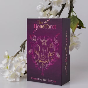 May include: A tarot card box with a dark purple background and the words "The Bone Tarot" in pink and gold. The box features a gold and pink illustration of a skull with floral accents. The text "Created by Sam Sawyer" is at the bottom.
