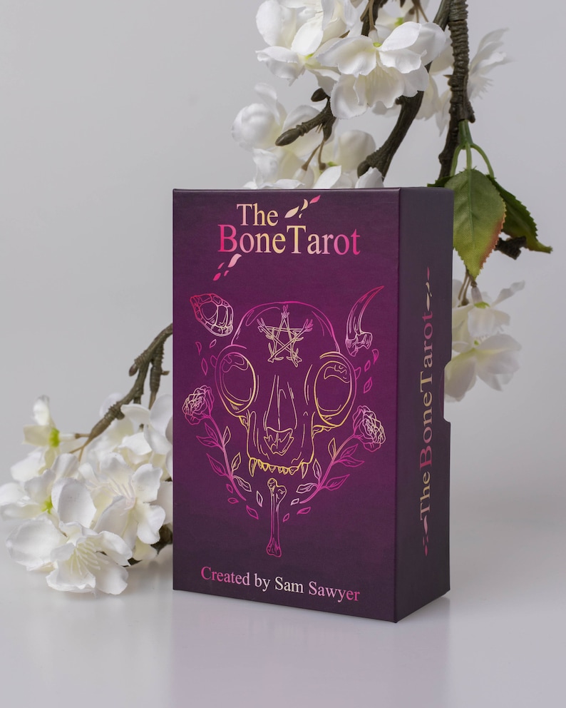 May include: A tarot card box with a dark purple background and the words "The Bone Tarot" in pink and gold. The box features a gold and pink illustration of a skull with floral accents. The text "Created by Sam Sawyer" is at the bottom.
