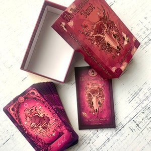 May include: A deck of tarot cards with a purple and pink color scheme. The box features a skull with antlers and floral accents. The text on the box reads "The Fae and the Ferns Tarot".
