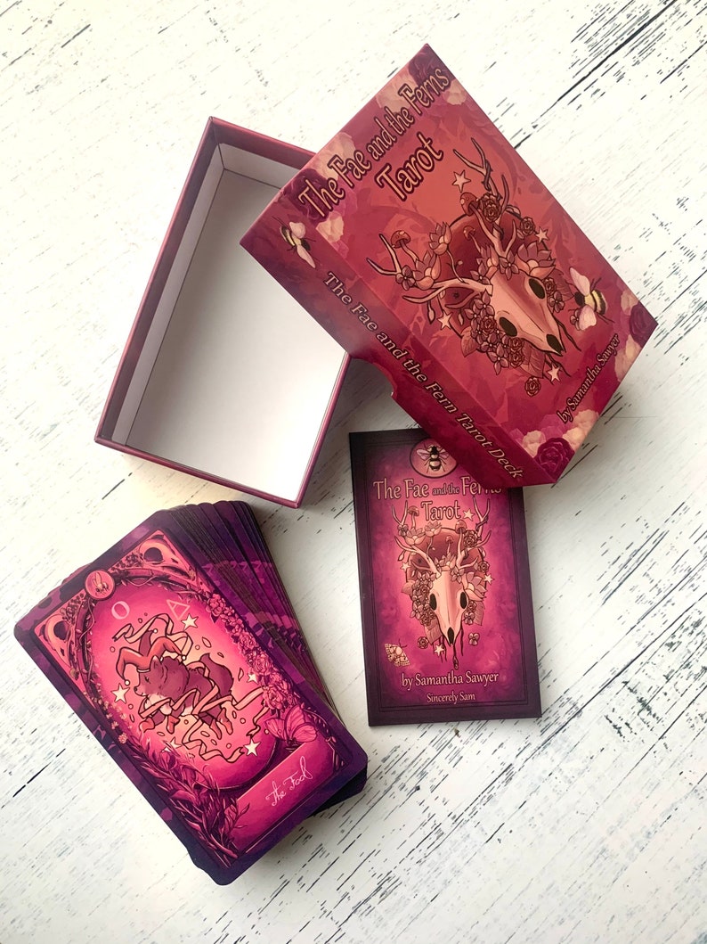 May include: A deck of tarot cards with a purple and pink color scheme. The box features a skull with antlers and floral accents. The text on the box reads "The Fae and the Ferns Tarot".