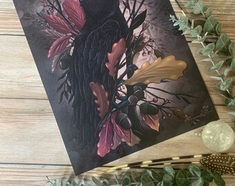 Puede incluir: Un cuervo negro posado en una rama con hojas de otoño en tonos burdeos, dorado y marrón. El cuervo se recorta sobre un fondo oscuro.