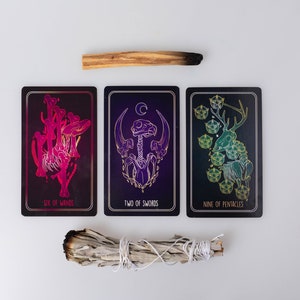 May include: Three tarot cards with intricate illustrations and text, including "Six of Wands," "Two of Swords," and "Nine of Pentacles." A bundle of white sage and a stick of palo santo are also present. The cards feature dark backgrounds with gold and vibrant color accents.