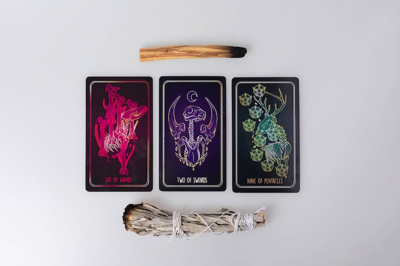 May include: Three tarot cards with intricate illustrations and text, including "Six of Wands," "Two of Swords," and "Nine of Pentacles." A bundle of white sage and a stick of palo santo are also present. The cards feature dark backgrounds with gold and vibrant color accents.