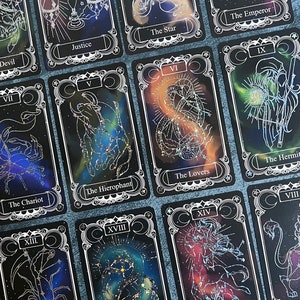May include: A collection of rectangular tarot cards featuring black backgrounds with colorful nebula-like patterns and silver foil line art illustrations of constellations and symbols. Each card displays a Roman numeral and a title such as Justice, The Star, The Emperor, The Hierophant, The Lovers, The Hermit, The Chariot, Death, The Moon, and Temperance.