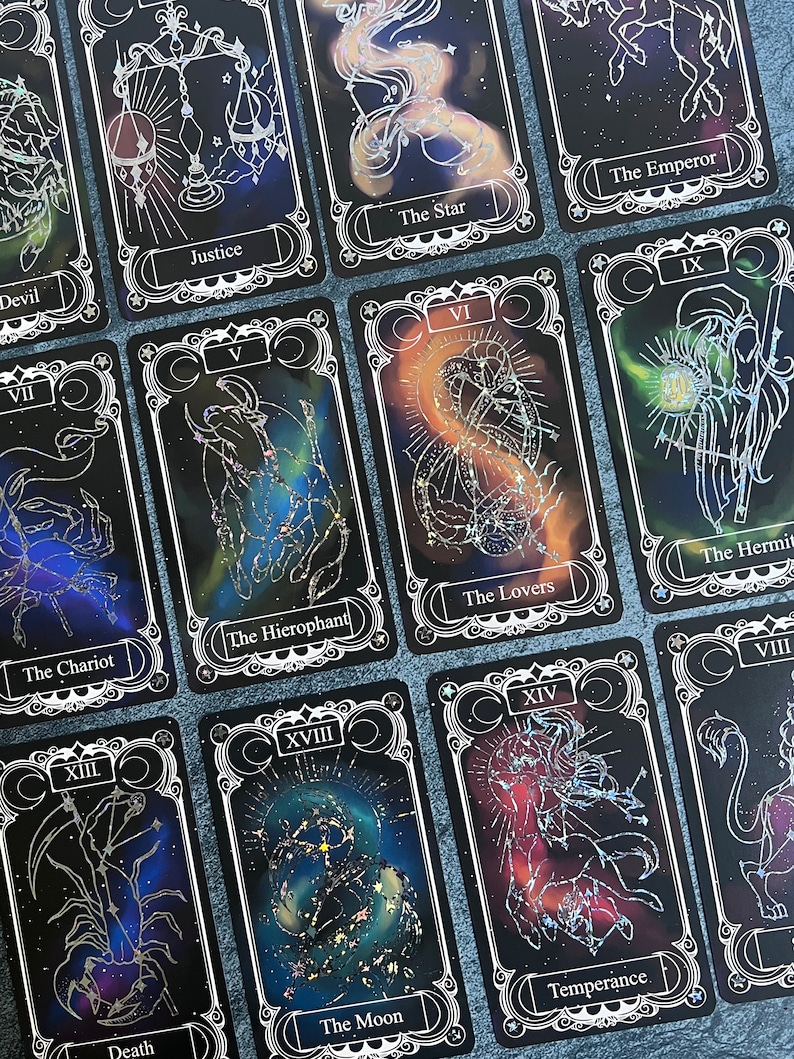 May include: A collection of rectangular tarot cards featuring black backgrounds with colorful nebula-like patterns and silver foil line art illustrations of constellations and symbols. Each card displays a Roman numeral and a title such as Justice, The Star, The Emperor, The Hierophant, The Lovers, The Hermit, The Chariot, Death, The Moon, and Temperance.
