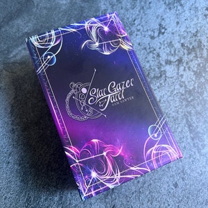 May include: A rectangular deck of Star Gazer Tarot cards by Sam Sawyer featuring a deep purple and blue cosmic background with white celestial line art, glowing nebula patterns, and a central logo with a crescent moon and star constellation design.