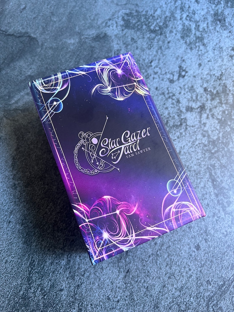 May include: A rectangular deck of Star Gazer Tarot cards by Sam Sawyer featuring a deep purple and blue cosmic background with white celestial line art, glowing nebula patterns, and a central logo with a crescent moon and star constellation design.