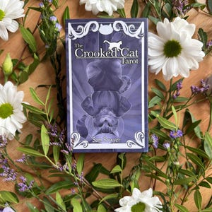 May include: A rectangular tarot deck box with a purple and white color scheme. The front features an illustration of a cat hanging upside down with the text The Crooked Cat Tarot and the author name Sam Sawyer printed in white lettering.