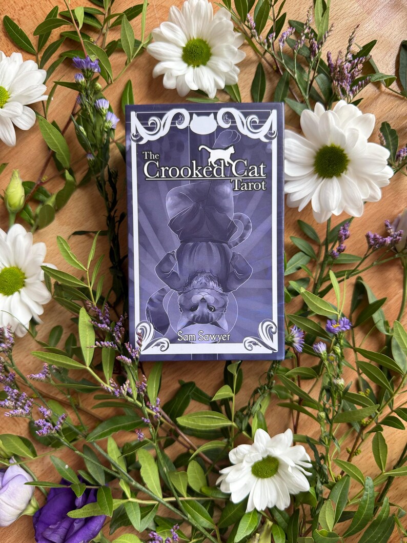 May include: A rectangular tarot deck box with a purple and white color scheme. The front features an illustration of a cat hanging upside down with the text The Crooked Cat Tarot and the author name Sam Sawyer printed in white lettering.