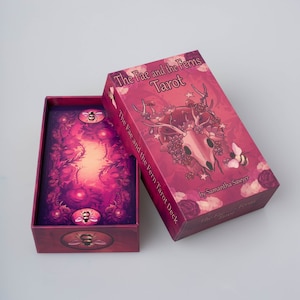 May include: A closed box of The Fae and the Ferns Tarot deck. The box is red with a pink and purple illustration of a deer skull with flowers and leaves. The text "The Fae and the Ferns Tarot" is printed in white on the box. The text "The Fae and the Fern Tarot Deck" is printed in white on the side of the box. The text "by Samantha Sawyer" is printed in white on the bottom of the box.